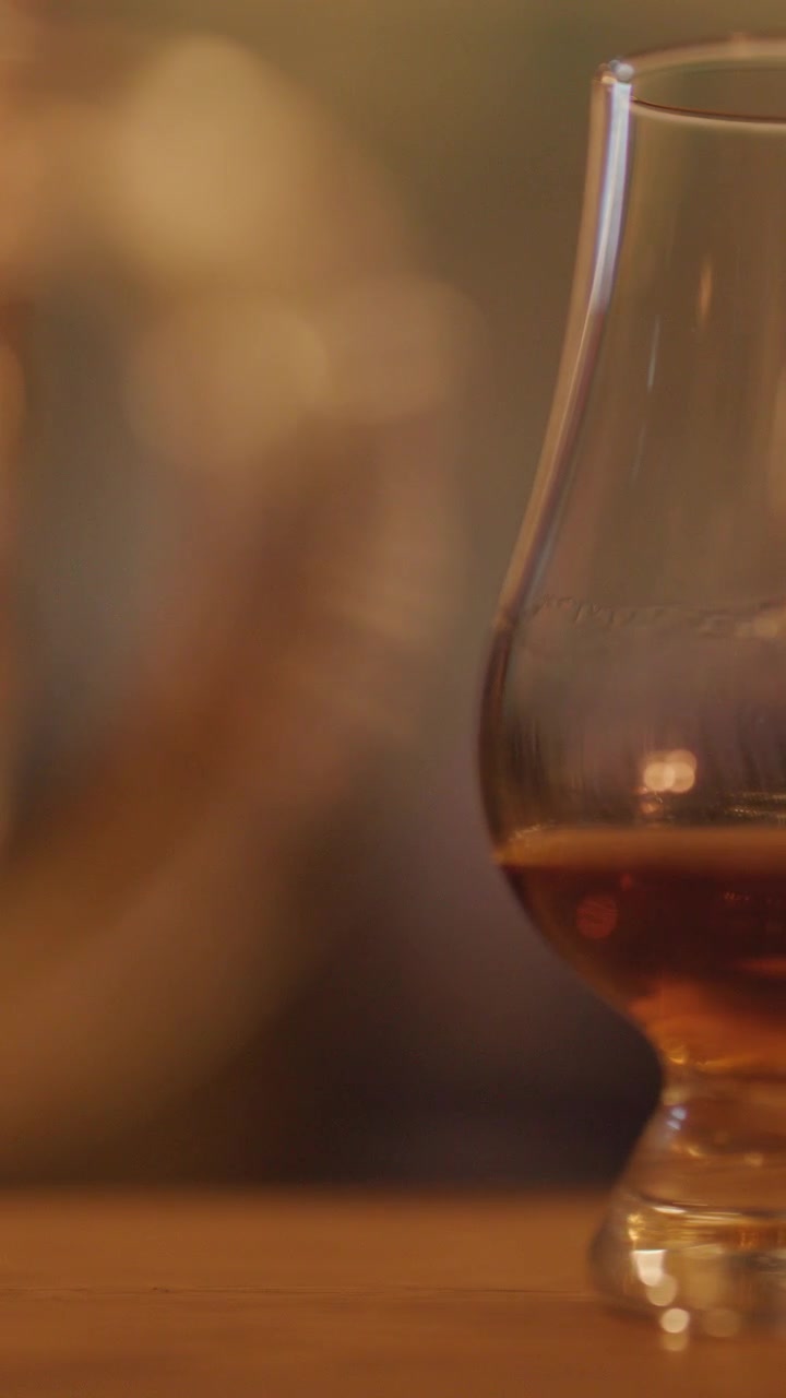 Un bicchiere da whisky con liquido ambrato poggia su un tavolo di legno, sfumando gradualmente mentre la calda luce di una candela crea uno sfondo bokeh morbido e suggestivo.