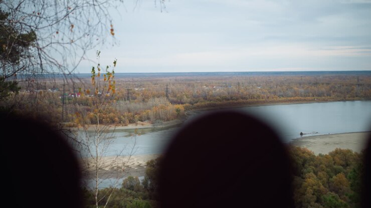 Scenic nature view featuring colorful autumn trees along a riverbank, vibrant fall foliage reflecting in peaceful river, with distant views of rolling landscapes under overcast sky