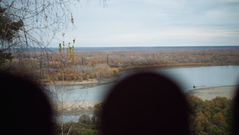 scenic nature view featuring colorful autumn trees along a riverbank, vibrant fall foliage reflecting in peaceful river, with distant views of rolling landscapes under overcast sky