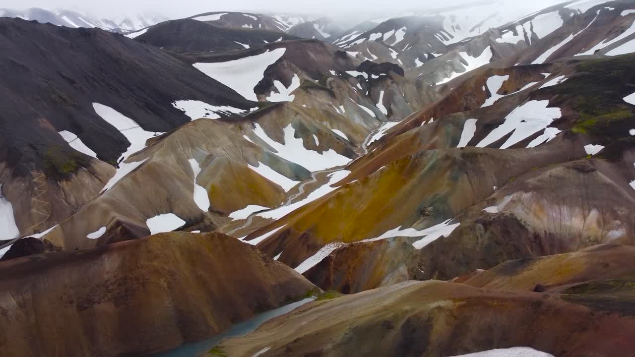 Aerial view of large volcanic terrain and brown colored Iceland landscape mountains during a cloudy day with white snow covering some parts of the steep and high altitude slopes. Misty clouds at back.