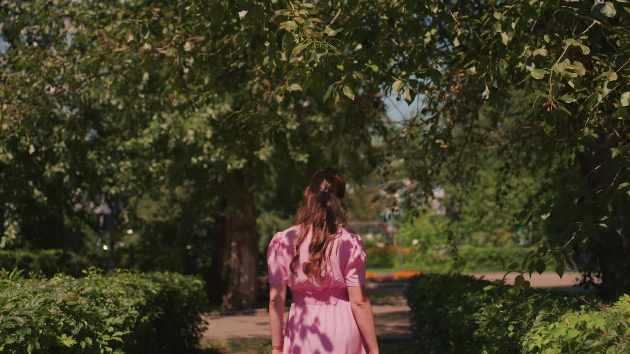 Mulher caucasiana caminhando por um caminho iluminado pelo sol com um vestido rosa, emoldurada por arbustos e árvores altas, ritmo lento e sereno, luz salpicada e bokeh suave, clima cinematográfico de verão, elegante retrato de moda