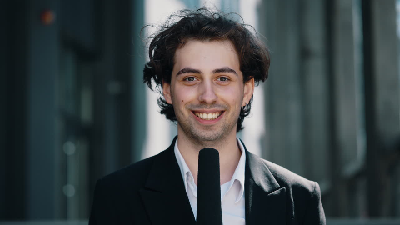 A young male reporter or journalist speaks into a microphone during an interview in an urban setting