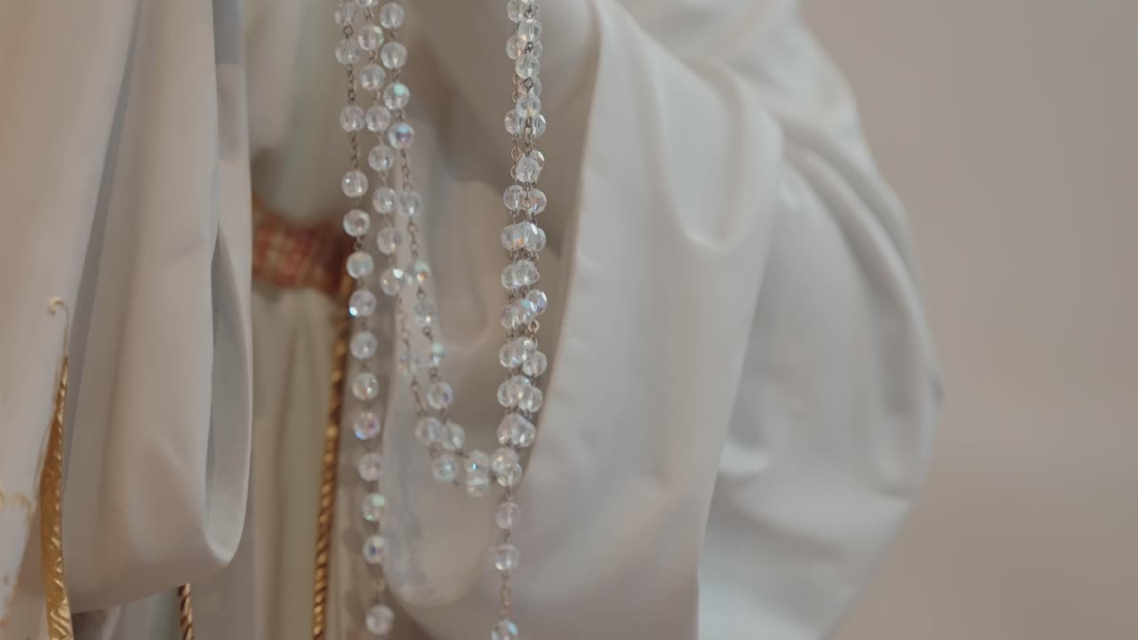sequence tilt of Virgin Mary statue holding crystal rosary beads, wearing a white and gold robe inside a softly lit church