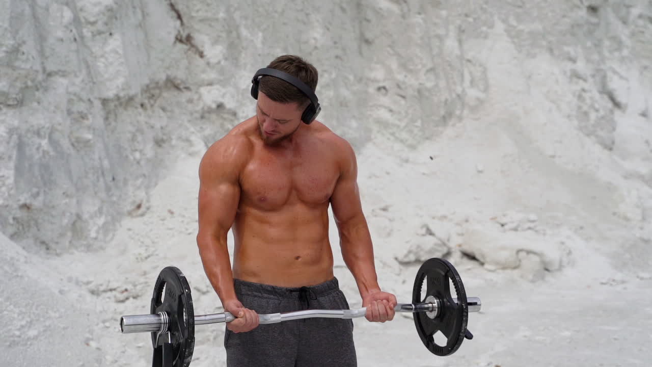 Concentrated muscular man exercising with heavy barbell in the mountains. Strong shirtless guy with headphones is lifting barbell. Young man training his muscles outdoors.