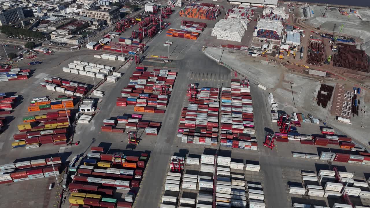 High-angle drone shot of Montevideo’s bustling container terminal, with cranes operating between stacks of colorful cargo containers by the waterfront