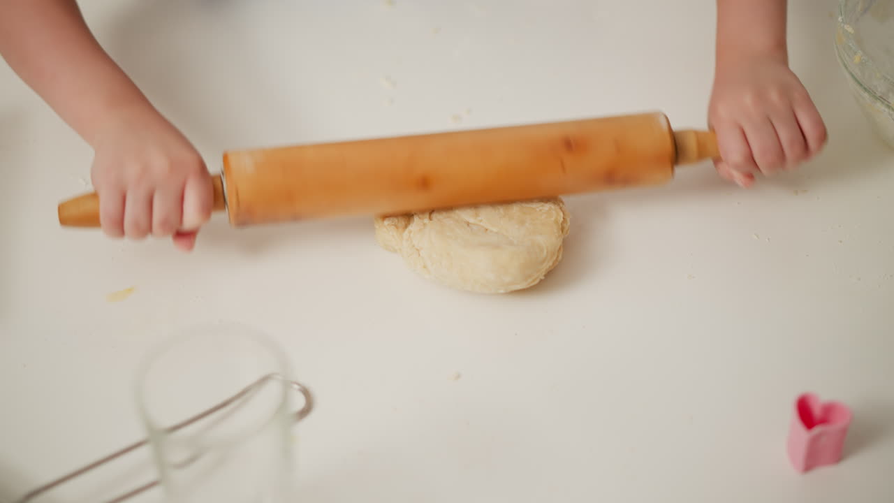 Kid rolling dough on white kitchen table during baking process using wooden roller pin with baking tools scattered around, preparing soft dough for homemade pastry in cozy bright kitchen environment