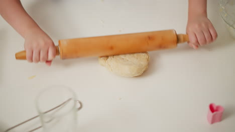 kid rolling dough on white kitchen table during baking process using wooden roller pin with baking tools scattered around, preparing soft dough for homemade pastry in cozy bright kitchen environment
