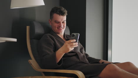 un hombre feliz y relajado está chateando en línea por teléfono inteligente enviando mensajes y sonriendo sentado en una tumbona