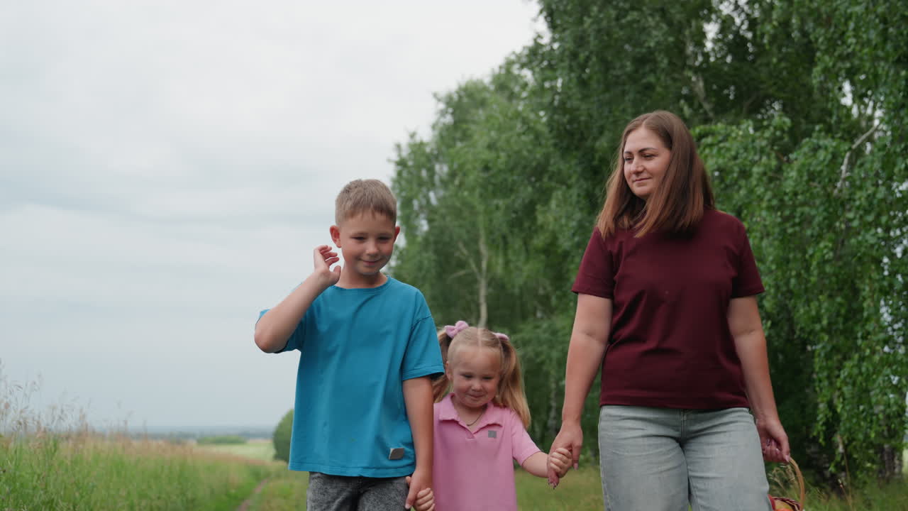 흰색 옷을 입은 엄마와 아이들이 손을 잡고 미소를 지으며 자작나무 밭의 초원을 걷고 있어요. 편안한 여름옷을 입었고 하늘은 흐립니다. 자연스러운 가족 간의 유대감을 보여주는 육아 관련 저속 촬영 영상입니다.