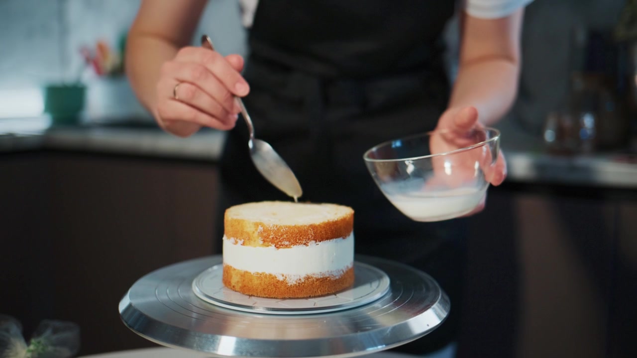 Person carefully spreads creamy frosting on a layered cake. The baker, wearing an apron, focuses intently on the task. Their hands move precisely, ensuring even coverage