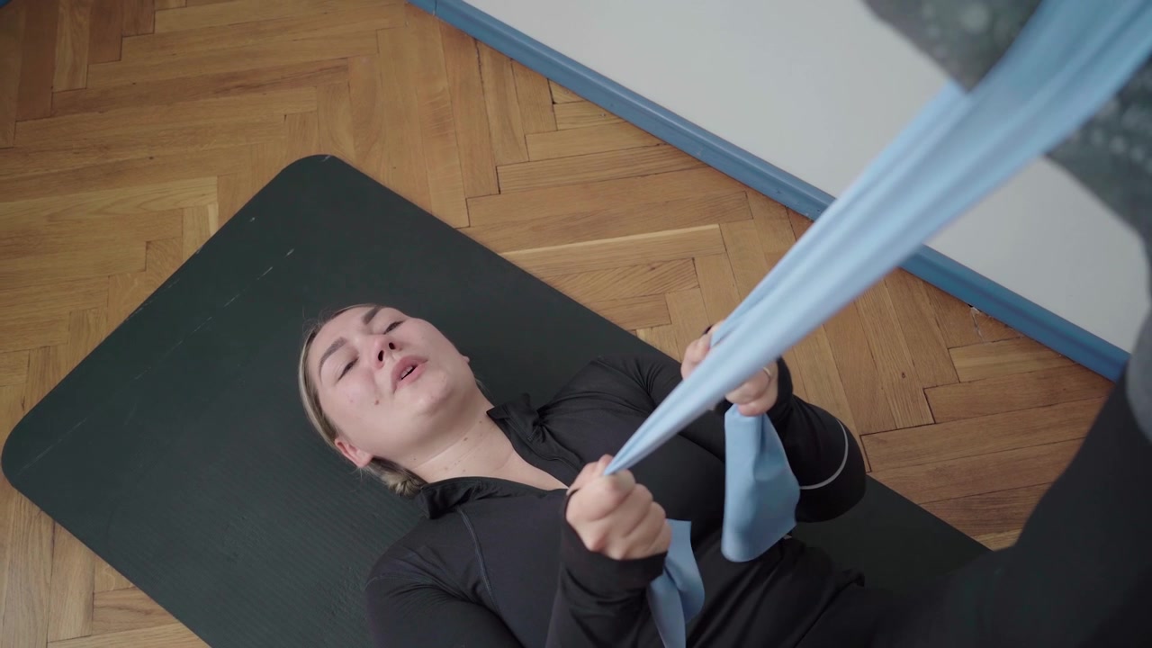 Pessoa plus size se exercitando deitada em um tapete, puxando uma faixa de resistência leve para um alongamento assistido dos isquiotibiais e exercício de reabilitação da perna