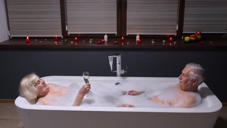 senior couple in love lying in warm bath with bubbles, enjoying relaxation, drinking champagne