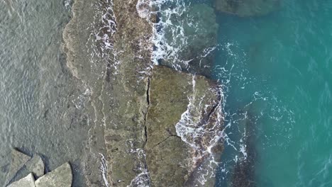 top down aerial view of protruding coastline rocks with waves crashing in turquoise ocean waters