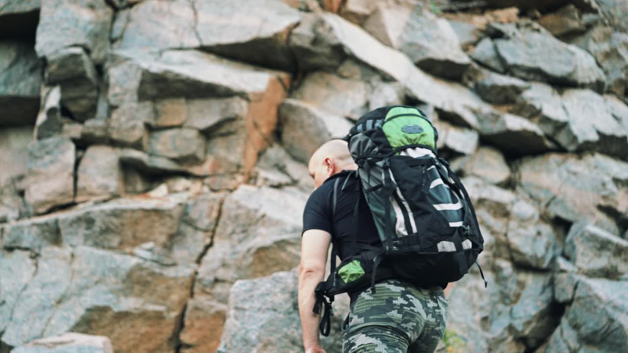 adult traveler in shorts and a t-shirt with a backpack on his shoulders is climbing rocks and examining locality around himself