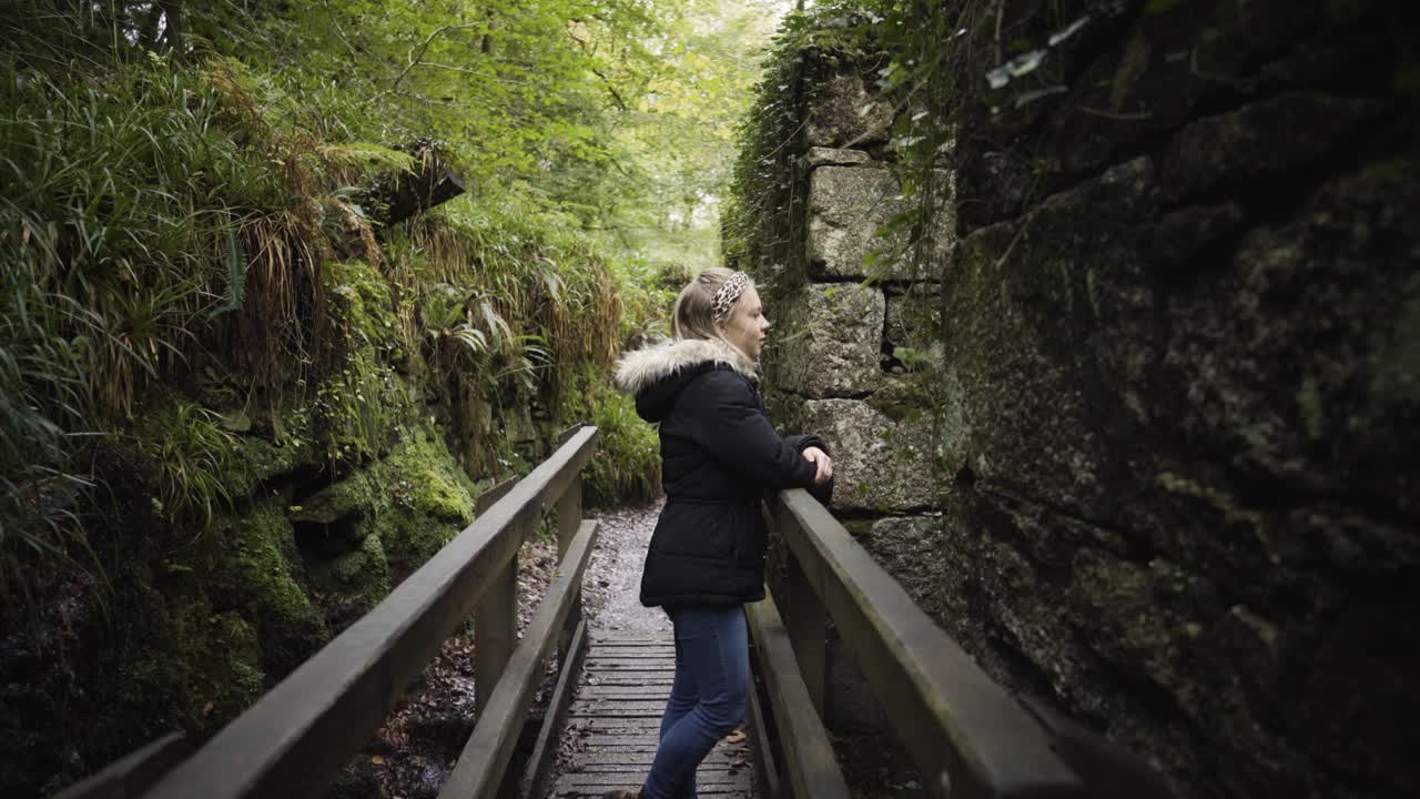 mujer joven de pie sobre una pasarela de madera frente a la pared en ruinas de una fábrica abandonada en la reserva natural de kennall vale en cornualles