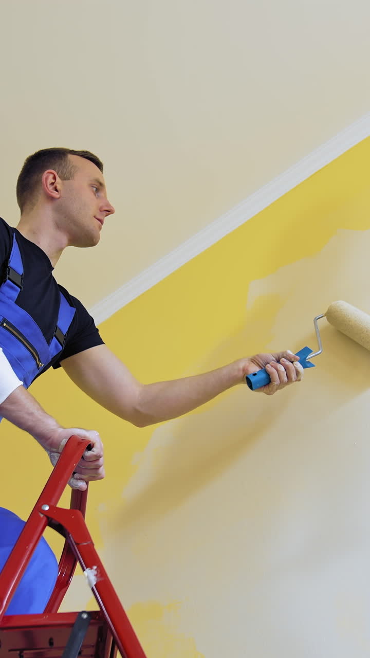 Professional craftsman painting walls indoors. Young male worker in blue overalls standing on a building ladder and use paint roller. View from below. Vertical video