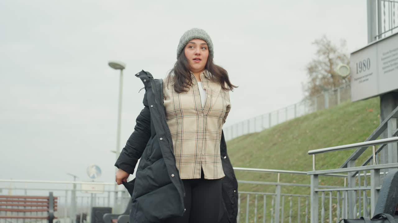 Wide shot of caucasian woman removing coat, shrugging jacket off to reveal knit sweater, overcast urban park with bench and railing, confident stride and playful sway, movement evokes dancelike