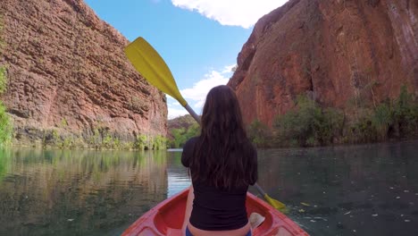 wide angle shot from behind of woman canoeing up a river in between large spectacular red rock cliffs