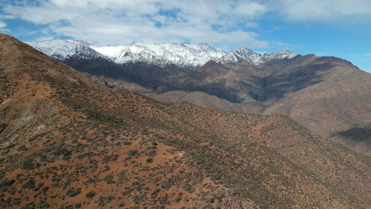 Veduta aerea con carrellata a destra di Morro Las Papas nel Parco San Carlos de Apoquindo, con una catena montuosa innevata sullo sfondo in una giornata limpida, Santiago, Cile