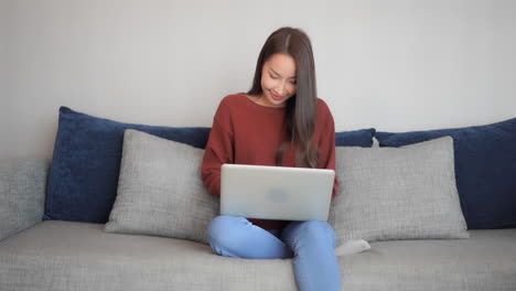 Mujer-Asiática-Sentada-En-El-Sofá-Con-Su-Laptop-En-El-Regazo-Y-Escribiendo-Que-Mostrando-Una-Expresión-Ganadora-Y-Levantando-La-Mano