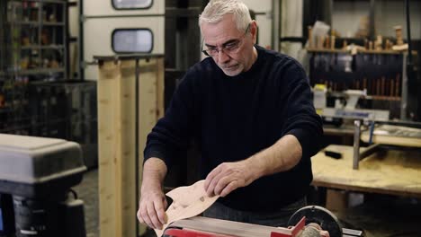 senior man in black sweater grinds the fish shape pattern with grinding machine with sawdust flying into the sides, profession, carpentry concept. caucasian man sanding wood at the workshop. slow motion