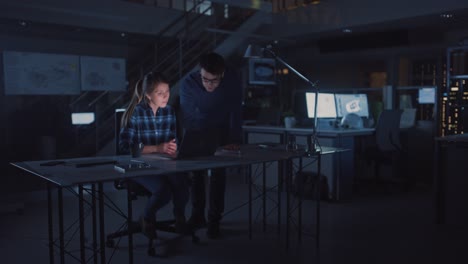 industrial engineering facility: female engineer working on desktop computer, project manager stands beside and explains specifics of the task and project details. working late on engine prototype