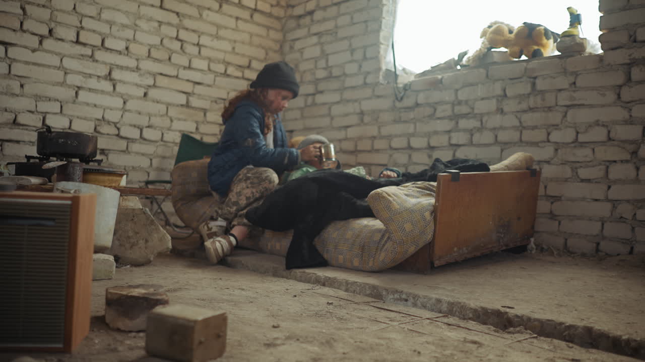 Girl in winter jacket and hat gently feeds sick friend lying on worn mattress inside cold abandoned brick shelter, surrounded by scattered old cooking pots, emotional care after apocalypse