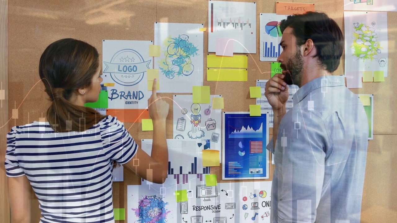 Uomo che prende una nota adesiva su una bacheca di sughero mentre una donna sistema grafici di brand per il marketing.
