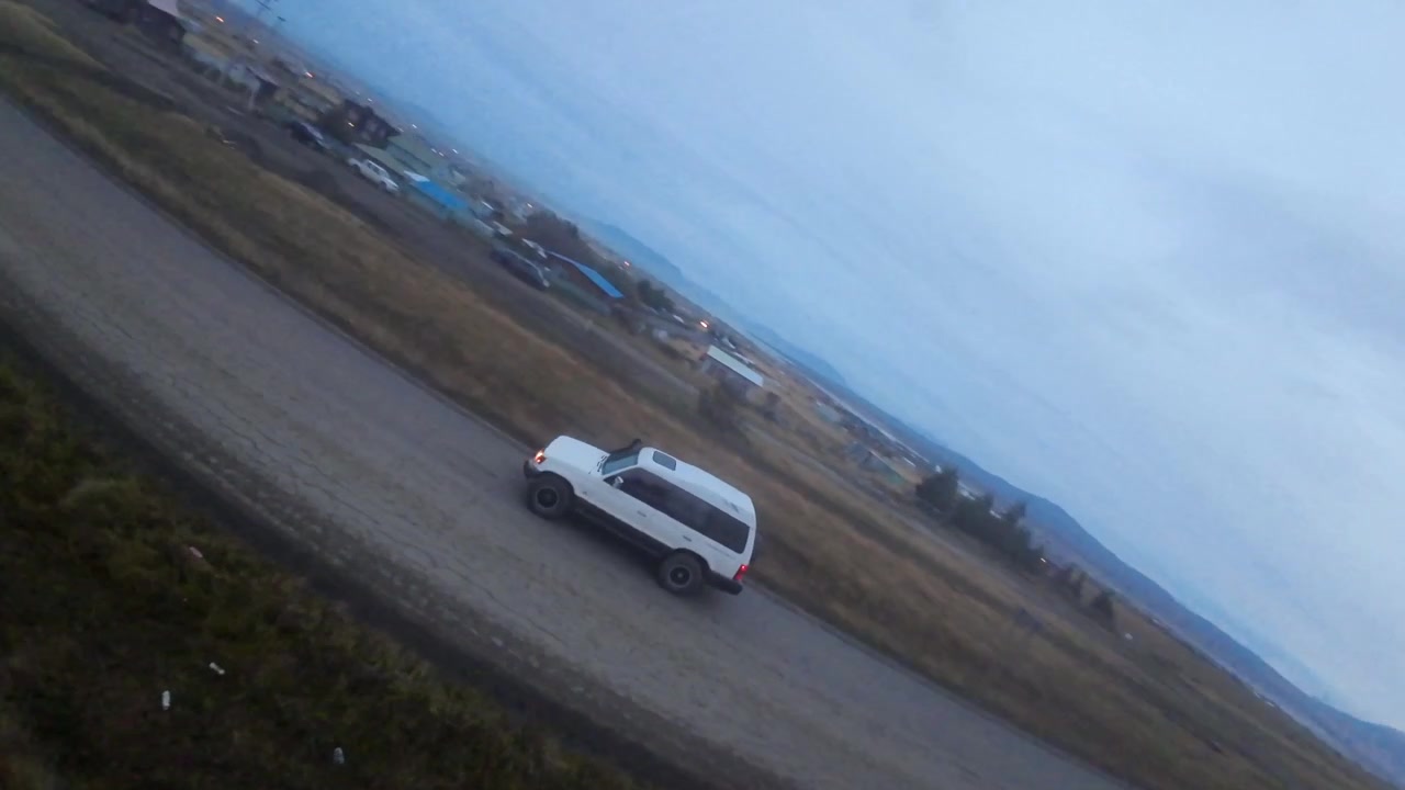 Um drone FPV voa ao lado de um veículo 4x4 branco percorrendo uma estrada de cascalho, com campos abertos e colinas perto de Puerto Natales, Chile
