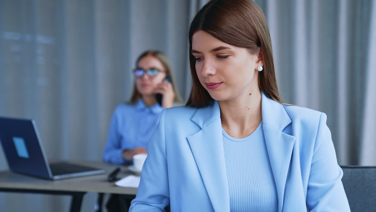 Lady office employee puts off her paper notebook and takes laptop. Working hours in office. Female colleague speaks on the phone at backdrop in blur.