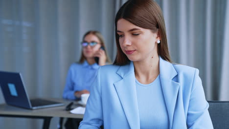 lady office employee puts off her paper notebook and takes laptop. working hours in office. female colleague speaks on the phone at backdrop in blur.