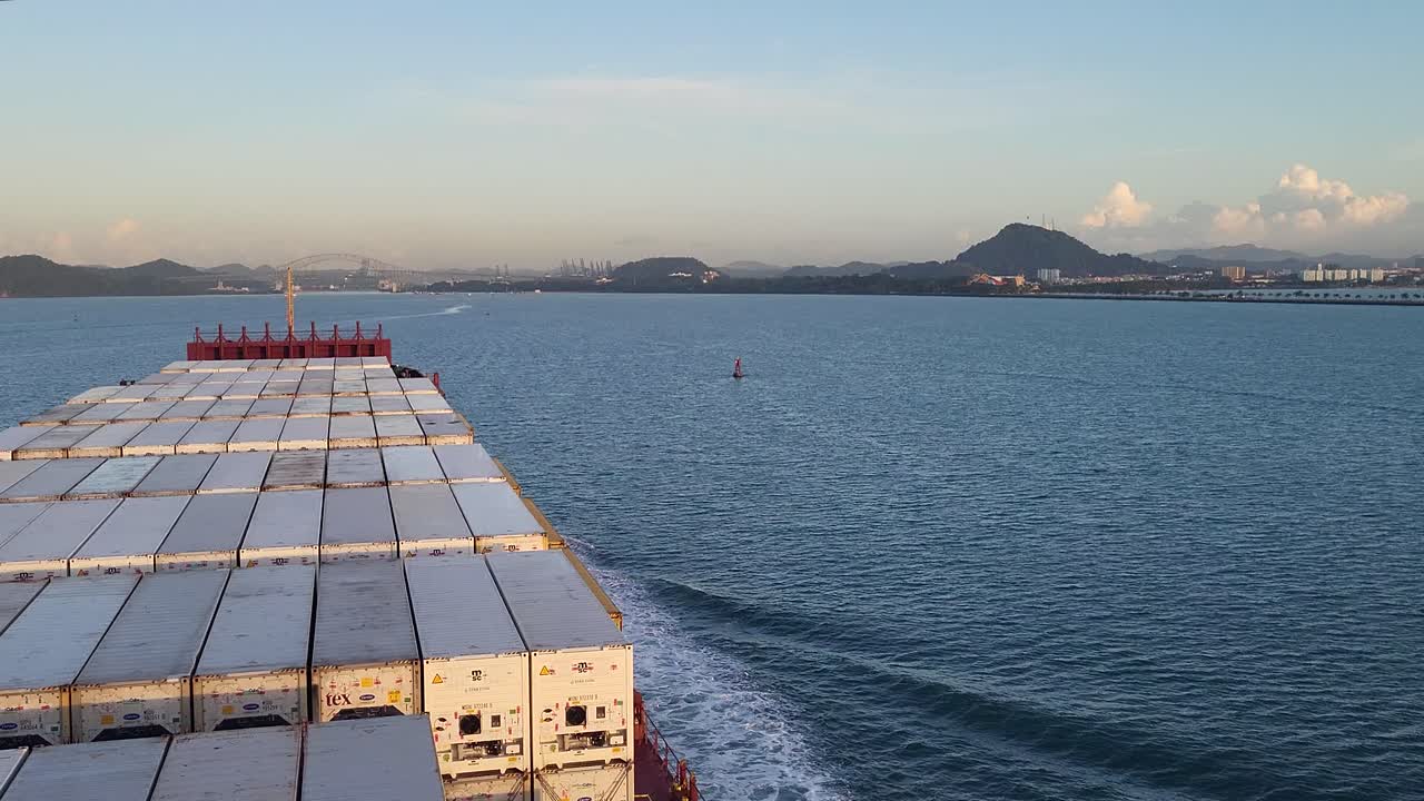 The view from aboard a massive container ship at dawn shows the vessel's bow cutting through the water of the Panama Canal as the sun rises over the distant coastline and city.