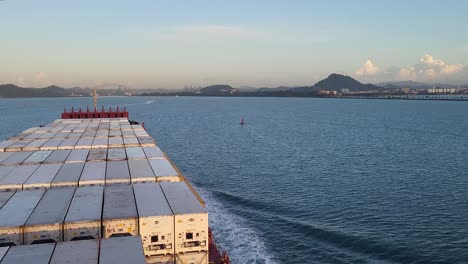 the view from aboard a massive container ship at dawn shows the vessel's bow cutting through the water of the panama canal as the sun rises over the distant coastline and city.