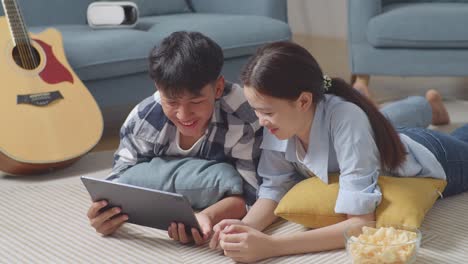 asian teen couple using tablet while lying on carpet on the floor at home. watching movie on tablet with snacks, speaking, and enjoying time together