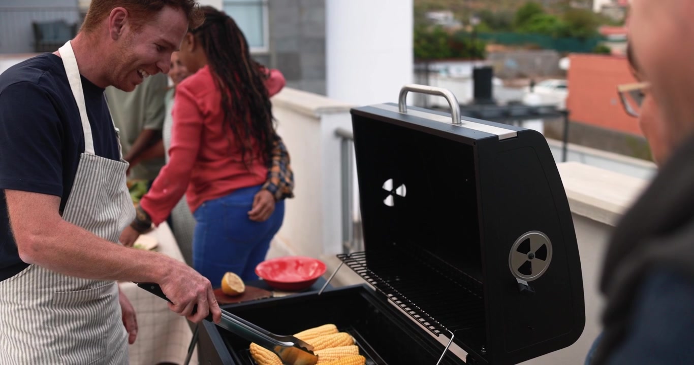 Uomo felice che griglia pannocchie al barbecue a una festa in terrazza