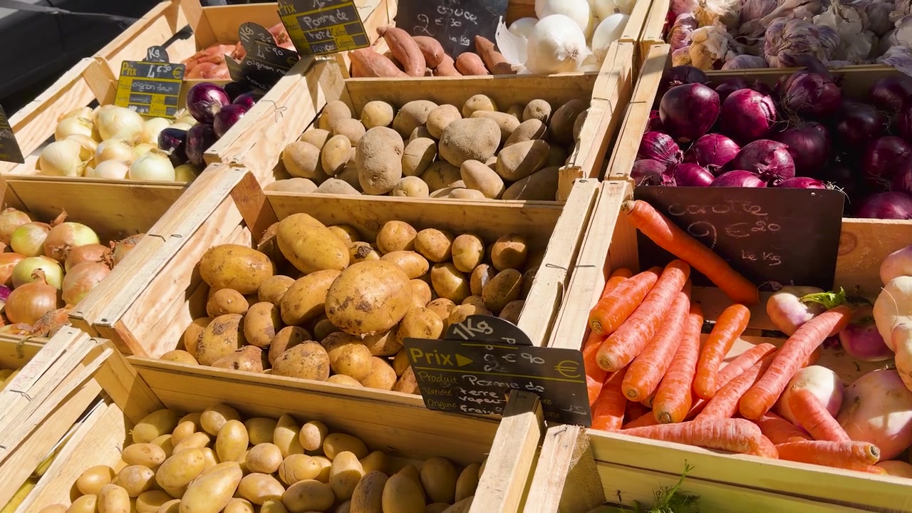 High angle panning shot of potatoes, onions, and carrots in wooden crates under bright sunlight