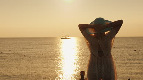 Una-Mujer-Joven-Con-Un-Vestido-Ligero-Y-Un-Sombrero-Se-Encuentra-Con-El-Amanecer-En-El-Mar,-Un-Barco-Es-Visible-En-La-Distancia-D