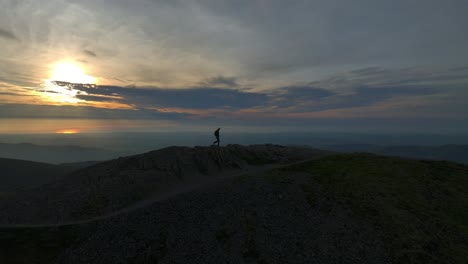 Einsamer-Bergwanderer,-Der-Sich-Während-Der-Goldenen-Stunde-Gegen-Den-Himmel-Mit-Der-Untergehenden-Sonne-Abhebt-Und-Den-Gipfelpfad-überquert-Und-Anhält