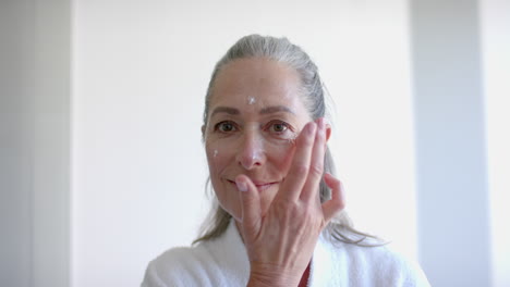applying face cream, mature woman in bathrobe caring for skin at home