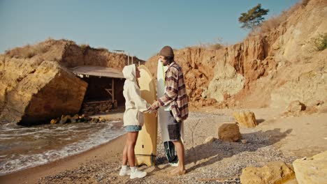 A-brunette-man-in-a-checkered-shirt-stands-with-his-blonde-girlfriend-in-a-white-sweatshirt,-looks-at-her-and-communicates,-they-hold-surfboards-in-their-hands-while-standing-on-a-sandy-rocky-beach-near-the-sea-in-the-morning