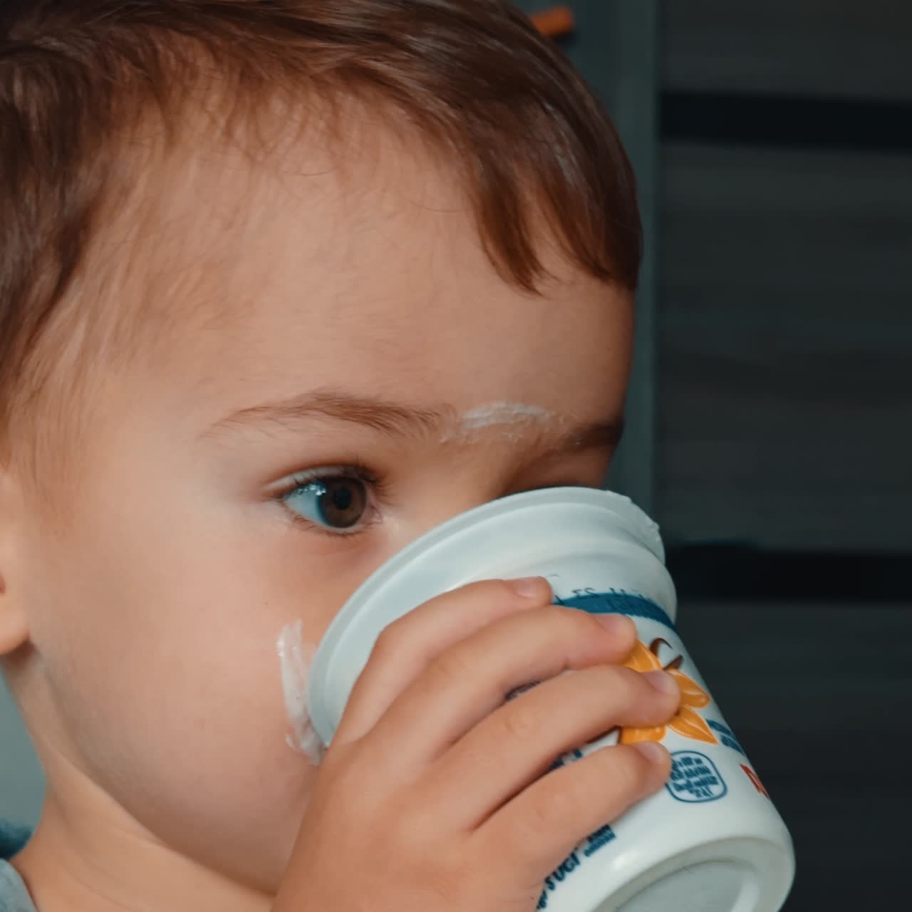 Bambino adorabile che mangia lo yogurt. Un bimbo carino usa un cucchiaio, beve da una tazza e ha macchie di latticini sul viso. Primo piano. Schermo verticale