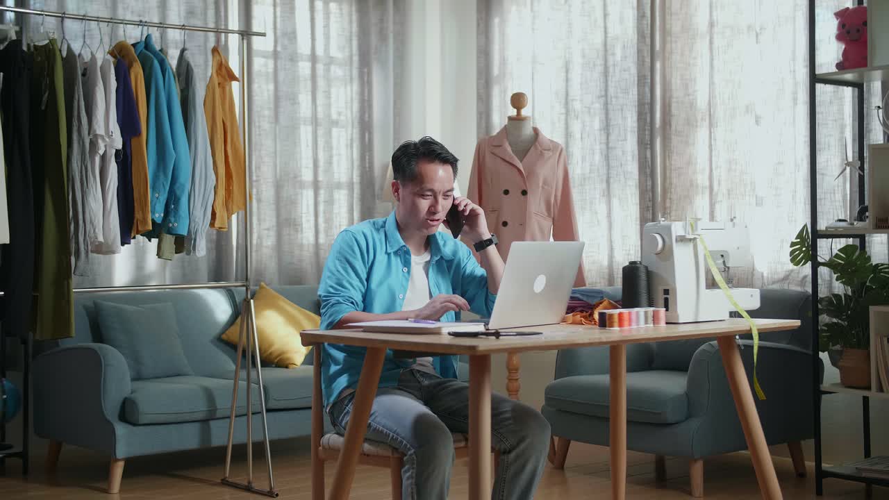 Asian Male Designer With Sewing Machine Talking On Smartphone While Designing Clothes On Laptop Computer In The Studio