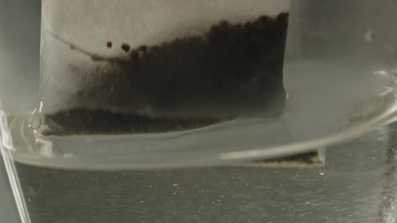 Macro of a tea bag being dropped in a glass of hot water in slow motion. The surface of the water bounces up and down while the teabag is being pulled