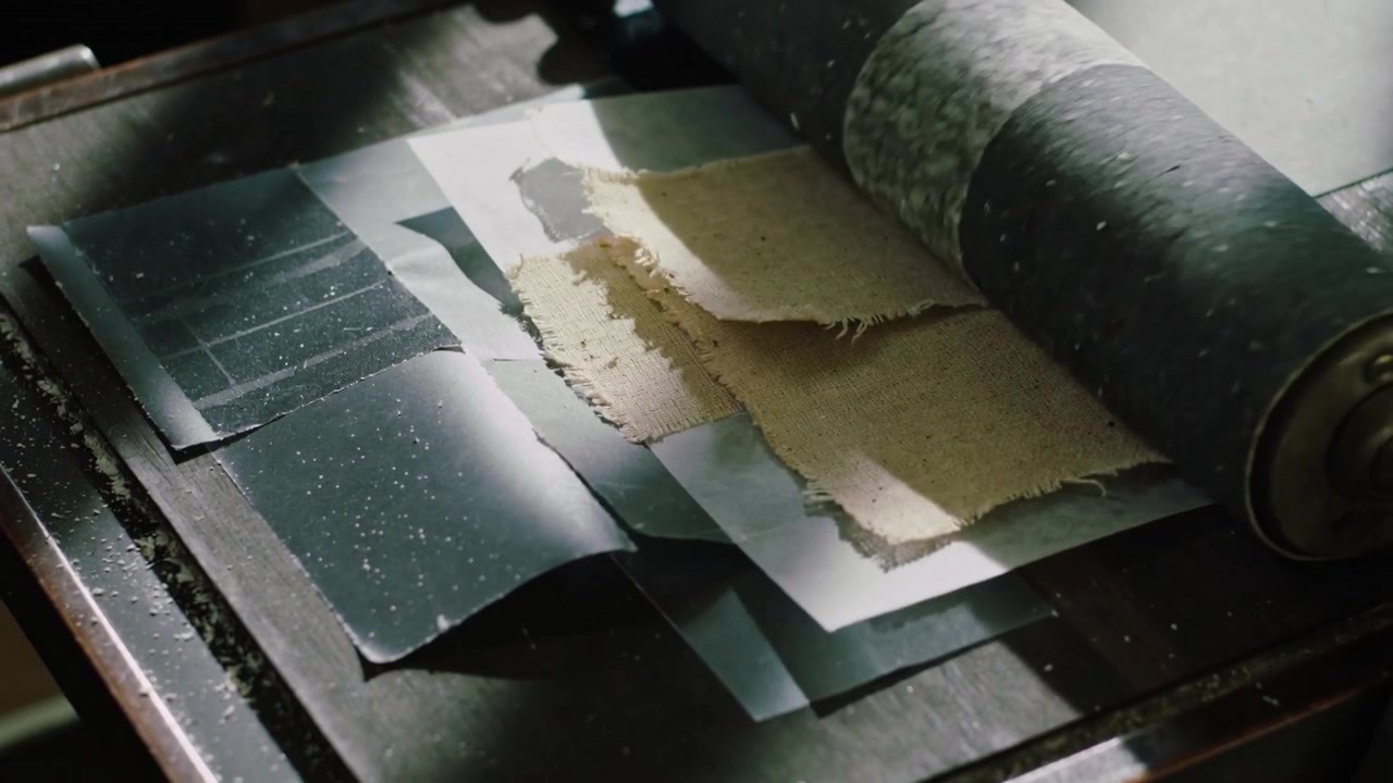 Descending roller advancing right, pressing white stack with torn tan fabric on table, flattening