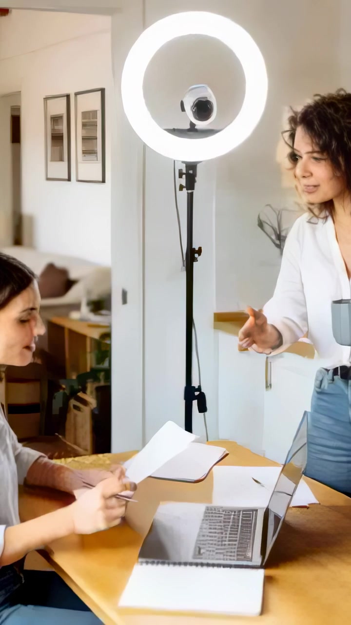 Vertical video: Right woman speaking, two women reviewing papers at home with ring light, in shirts