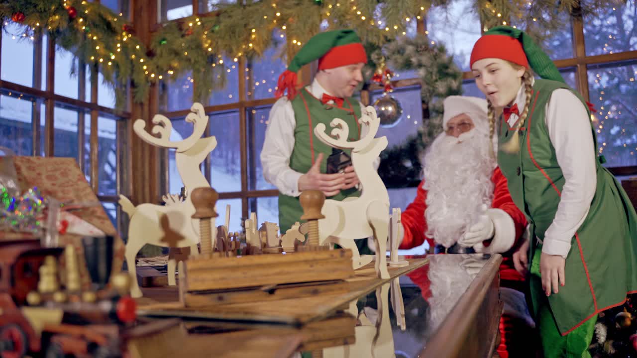 Wooden handmade toys on the table. Santa Claus and two elves with presents on Christmas background indoors.