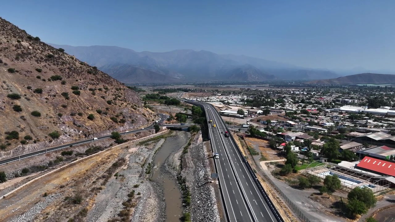 Ripresa aerea orbitale sopra un ponte autostradale che attraversa il letto asciutto del fiume Aconcagua ai piedi di un arido pendio montuoso, rivelando la città di Los Andes e le cime andine sullo sfondo. Los Andes, Cile.