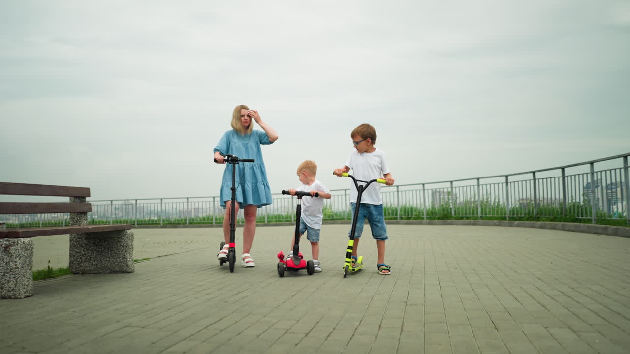 A mother and her two children ride scooters together in an open park area, with an iron rail by the side and benches around