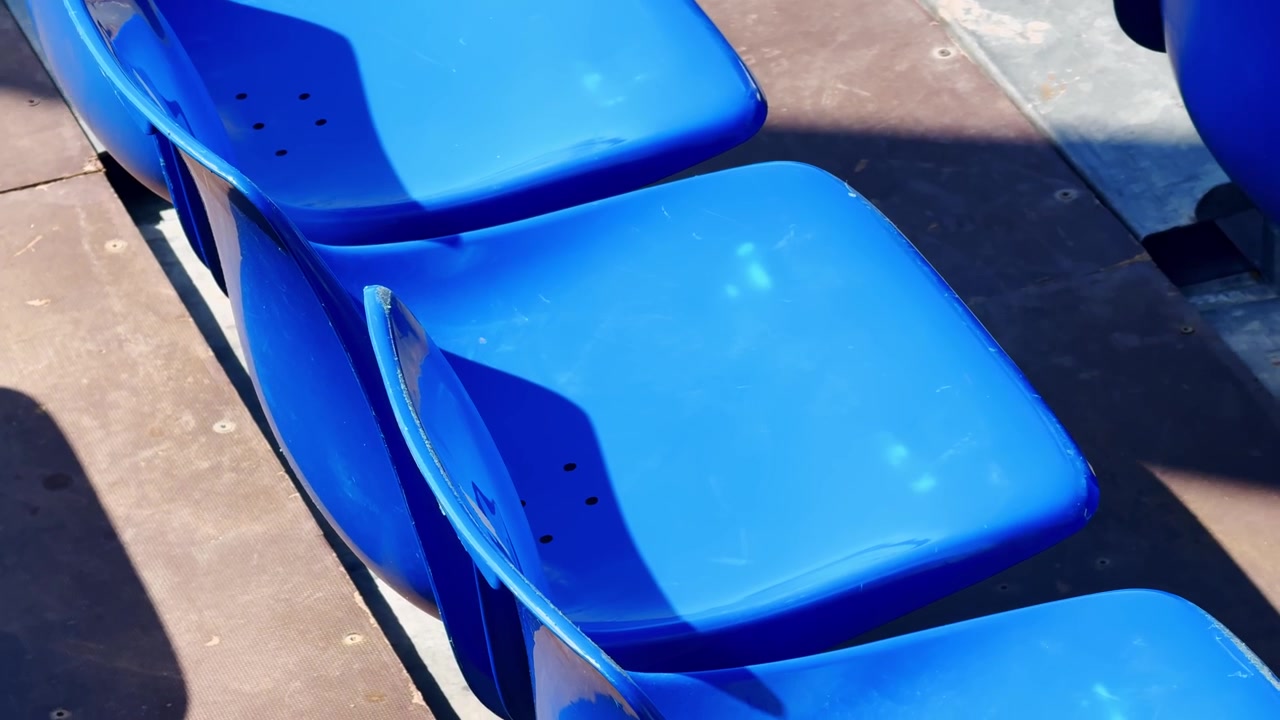 Primo piano di sedili da stadio in plastica blu brillante nelle tribune di un campo da tennis, con luce solare, ombre, gradini in metallo e dettagli dei posti a sedere vuoti per gli spettatori