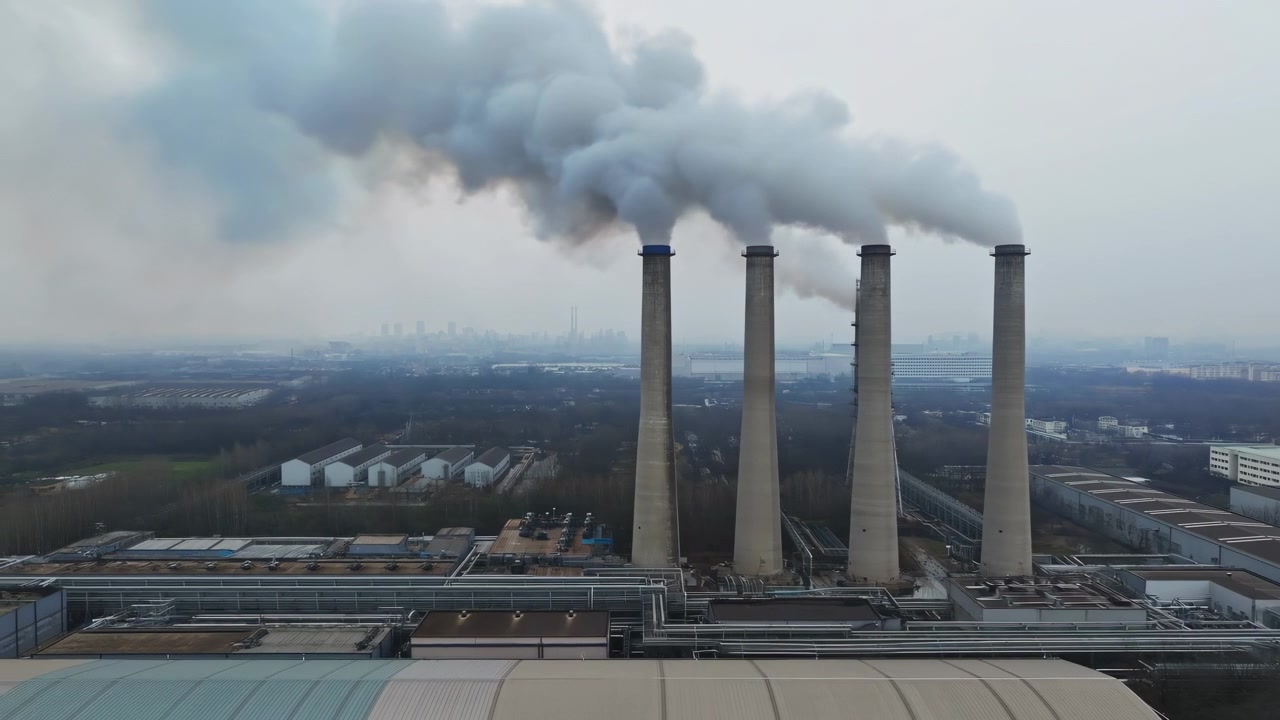 Vista molto elevata mostra quattro alte ciminiere di cemento che rilasciano attivamente fumo denso grigio chiaro che si diffonde attraverso il cielo coperto sopra i tetti industriali, creando una scena attenuata dell'industria energetica con